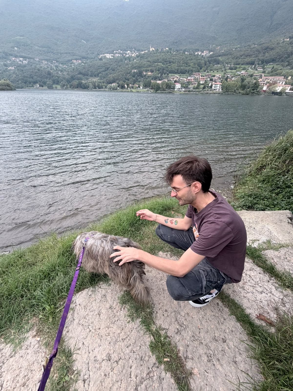 Gio Marco Baglioni accucciato sulla riva del lago con il suo cane, la Valle Camonica dietro