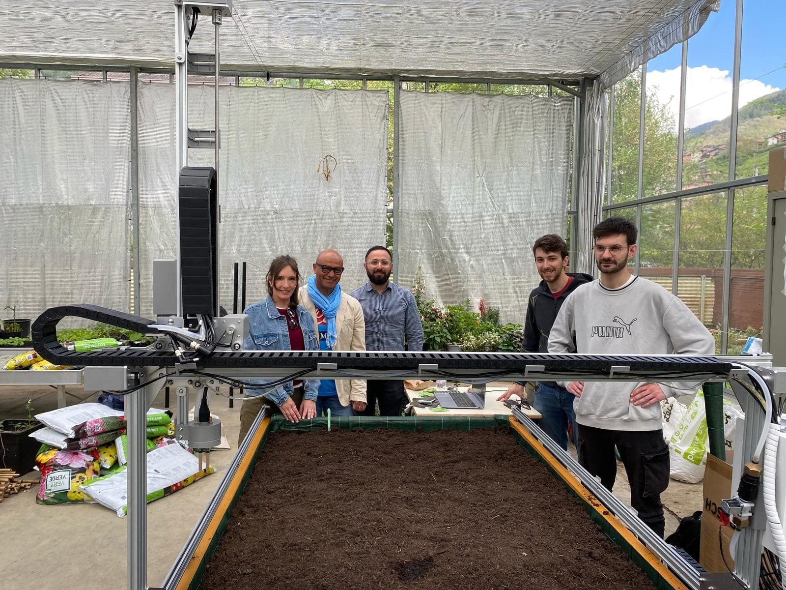 Cinque persone attorno al letto di coltivazione di un FarmBot, il robot da orto, dentro una serra